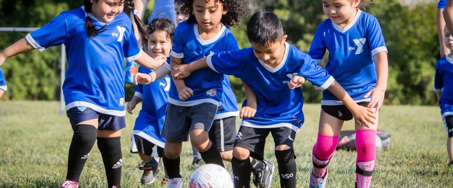 Group of children wearing YMCA soccer uniforms running after soccer ball